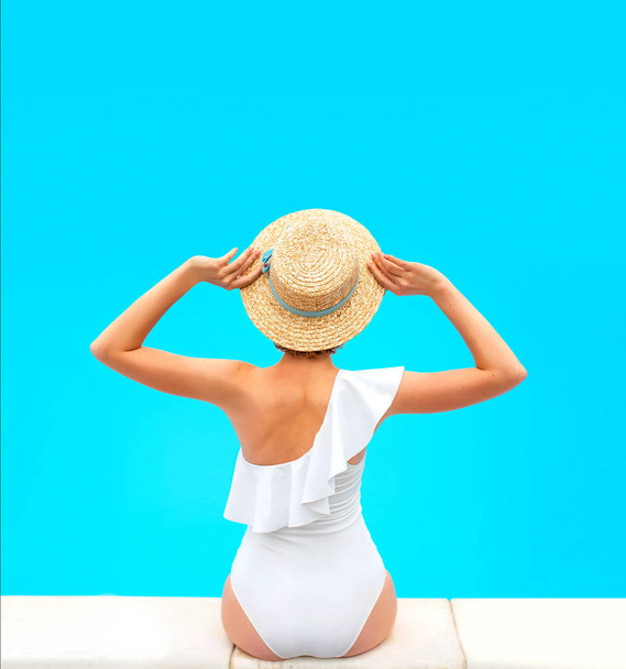 beautiful redhead (ginger) woman's back in the white swimminig suit sitting by the swimming pool (or sea or ocean) in the straw hat in summer day. Summer, relax, wellness, recreation concept - Fotó, kép