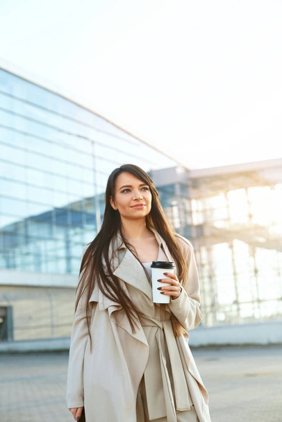 Business woman holding coffee and using phone near office. Portrait of beautiful smiling female with phone, standing outdoors. Phone communication.  - Foto, afbeelding