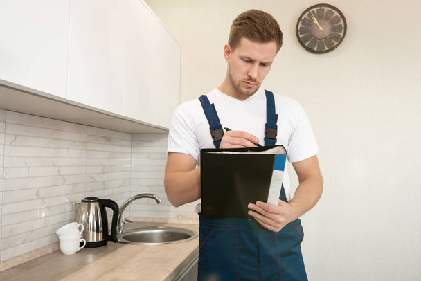 young handsome man plumber looks concentrated while filling guarantee papers after finishing installation work in the kitchen professional plumbing repair service - Фото, изображение
