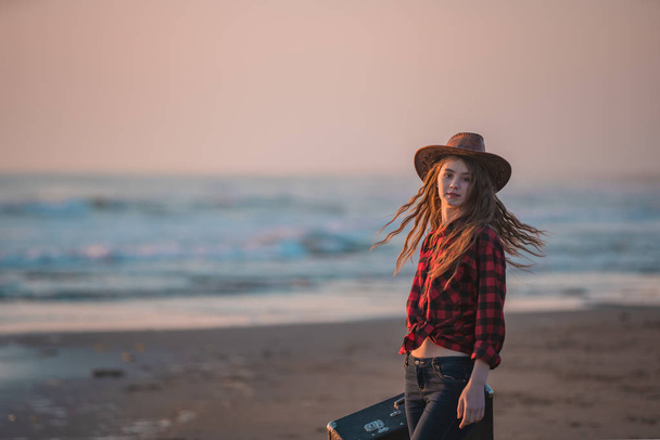 Mladý štíhlý dlouhosrstý bruneta v červených šatech kráčí bosý po tropické pláži s kufrem v ruce při západu slunce na den podél příboje na pozadí moře. - Fotografie, Obrázek