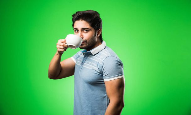 Hombre indio con taza de té / café o taza. Beber, presentar o sostener de pie aislado sobre fondo verde - Foto, Imagen