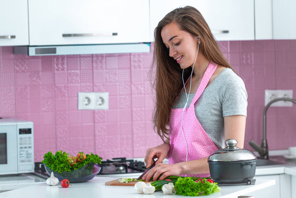 Young housewife in headphones and in an apron listening music and enjoying cooking preparation in kitchen at home. Love cooking. Clean healthy food and diet  - Фото, зображення