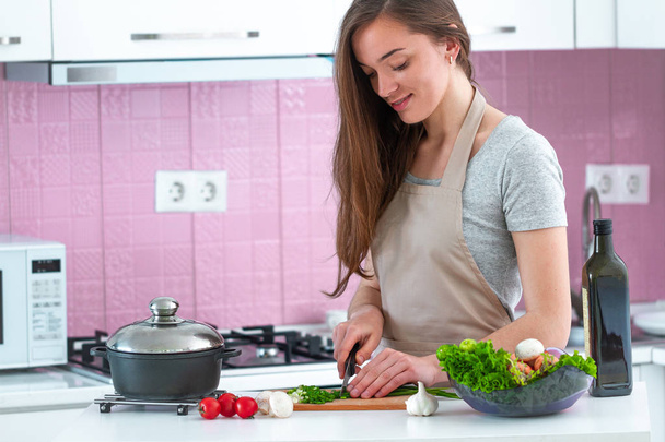 Atrakcyjna młoda kucharka w fartuchu siekająca warzywa na sałatkę na obiad w kuchni w domu. Czysta zdrowa żywność - Zdjęcie, obraz
