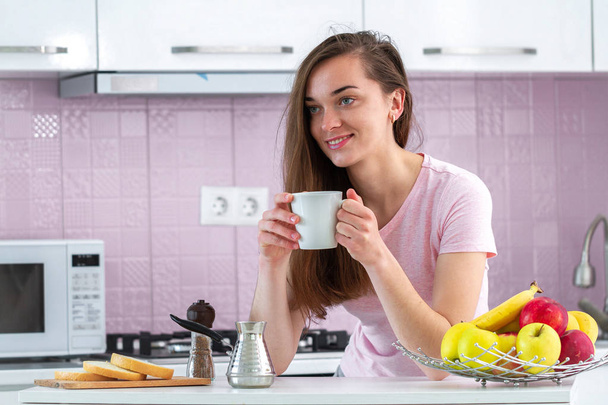 Feliz, atractiva, mujer bebe café caliente y sabroso para el desayuno por la mañana temprano en la cocina en casa. Buenos días y empezar un nuevo día
 - Foto, imagen