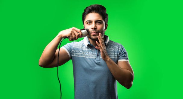 Indiase of Aziatische man scheerbaard met elektrische trimmer of Clipper staan geïsoleerd over groene achtergrond - Foto, afbeelding
