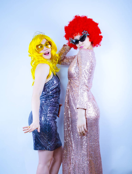 Two glamorous and beautiful girls in bright clothes and wigs have fun. Girlfriends posing while preparing for a Christmas party. Isolated on a light background. - Фото, изображение