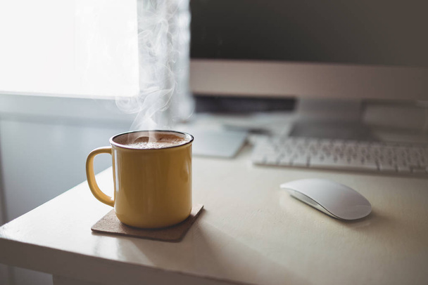 Uma xícara amarela de café fica na mesa do escritório sob a luz ensolarada da manhã à noite, ao lado do monitor e do teclado
 - Foto, Imagem