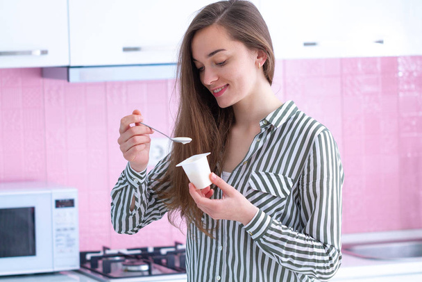 Portrait of a young, smiling, happy woman holding yogurt spoon and eating yogurt at home in the kitchen - Foto, immagini
