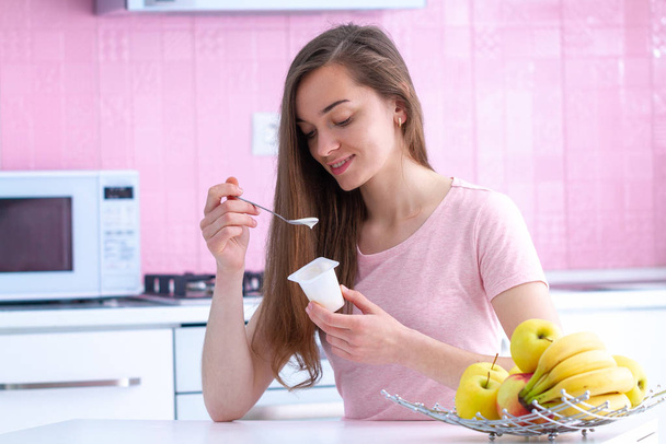 Νέα, ελκυστική μελαχρινή γυναίκα τρώει φρέσκο γιαούρτι για πρωινό νωρίς το πρωί στην κουζίνα στο σπίτι  - Φωτογραφία, εικόνα
