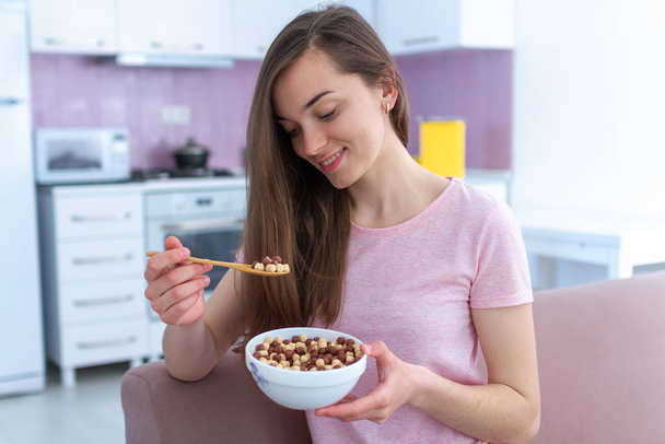 Portrait of young, happy beautiful brunette woman in pajamas eating chocolate balls for cereals breakfast at home  - Foto, imagen