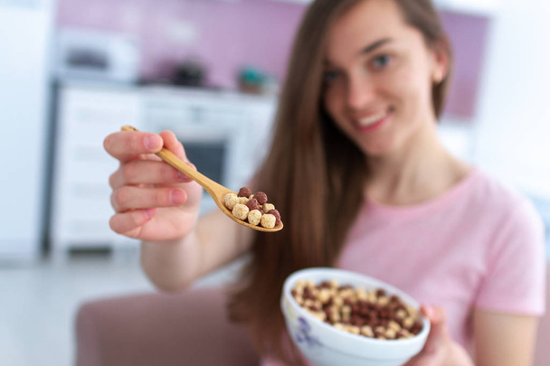 Young, smiling happy beautiful brunette woman holding cereals spoon and cereals bowl full of chocolate balls for breakfast at home - Фото, зображення