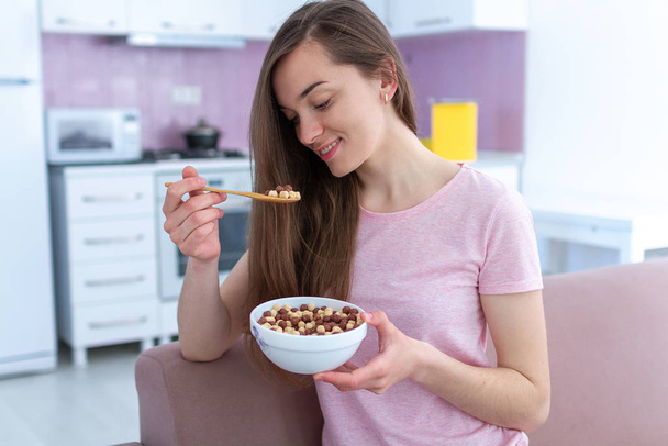 Portrait of young, happy beautiful brunette woman in pajamas eating chocolate balls for cereals breakfast at home  - Фото, изображение
