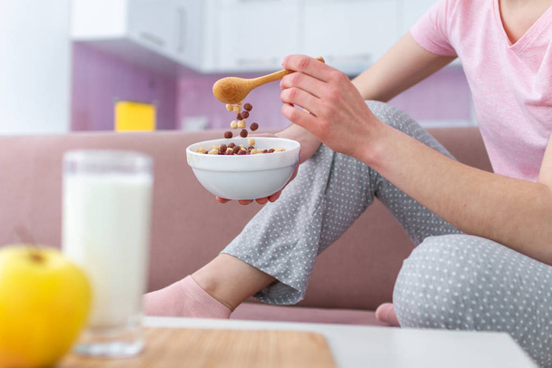 Woman in pajamas eating frosted chocolate balls, yellow apple and drinking fresh glass of milk for healthy  breakfast at home  - Photo, Image