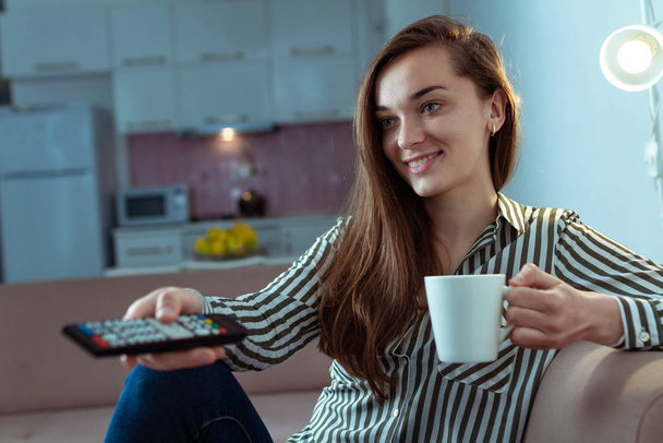 junge glücklich lächelnde Frau, die auf der Couch sitzt, sich ausruht und allein zu Hause verschiedene Fernsehprogramme anschaut  - Foto, Bild