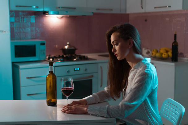 Lonely, unhappy, sad beautiful young woman in a blouse with glass of red wine is drinking alone in evening at home. Female alcoholism and alcohol addiction - Foto, immagini