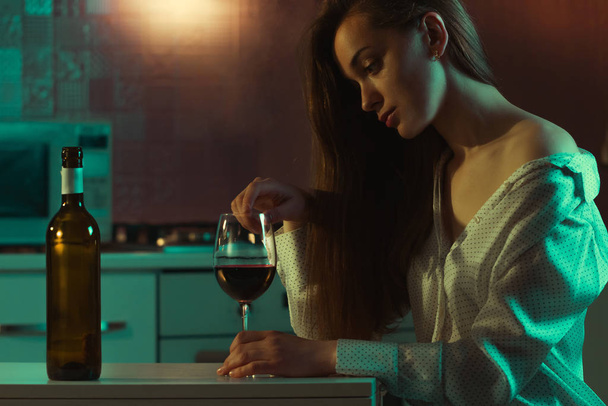 Lonely, sad, beautiful young woman with red wine is drinking alone at home in evening. Female alcoholism and alcohol addiction.  - Фото, зображення