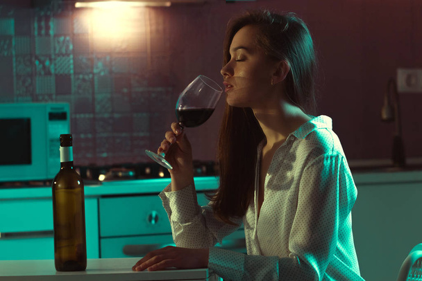 Lonely, beautiful young woman in a blouse with red wine is drinking in evening at home. Female alcoholism and alcohol addiction. - 写真・画像