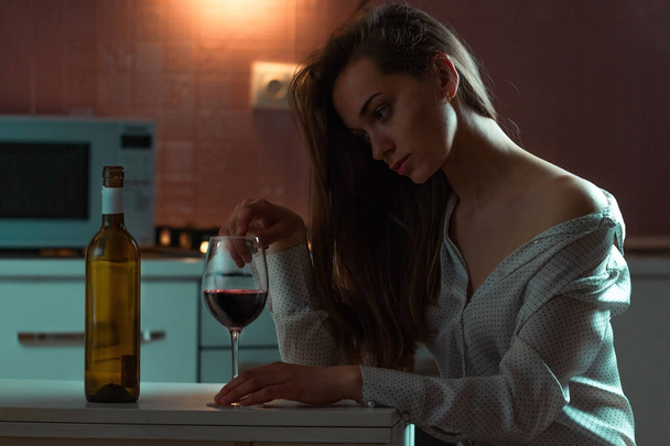 Lonely, sad beautiful young woman in a blouse with alcoholic beverage is drinking alone in evening at home. Female alcoholism and alcohol addiction - Фото, изображение