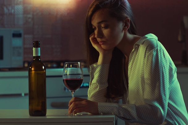 Lonely, sad, beautiful young woman with red wine is drinking alone in the kitchen at home in evening. Female alcoholism and alcohol addiction.  - Fotografie, Obrázek