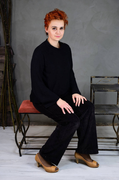 Art portrait of a pretty model with short red hair in dark clothes on a gray background in the interior. A woman sits on a chair right in front of the camera in various poses. - Foto, Imagen