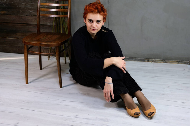 Art portrait of a pretty model with short red hair in dark clothes on a gray background in the interior. A woman sits next to a chair right in front of the camera in various poses. - Фото, изображение