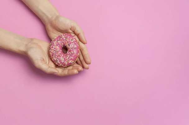 Красиві жіночі руки з ніжним манікюром тримають солодкий пончик на рожевому фоні зверху плоский простір для копіювання. Натуральні нігті, гелевий лак, догляд за собою, краса і мода. Догляд за нігтями. Тло пончика
 - Фото, зображення