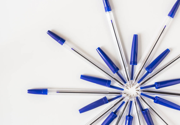 прозрачные шариковые ручки с голубой крышкой выкладываются в виде цветка на белом фоне
 - Фото, изображение