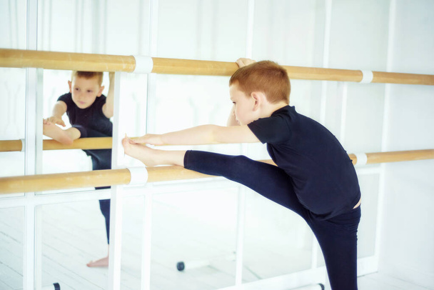 Junge Balletttänzer machen Übungen und dehnen sich auf Ballettstangen in der Nähe von Spiegeln im Ballettstudio mit weißem Licht. Tanzender Junge. Aktivitäten und Sport für Kinder. Harte Arbeit in der Kindheit, um Traum zu verwirklichen - Foto, Bild