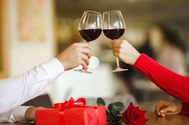 Hands holding glasses of wine on restaurant background. Couple clink glasses with red wine. Valentines day celebration concept. Relationship, surprise and love concept. - 写真・画像