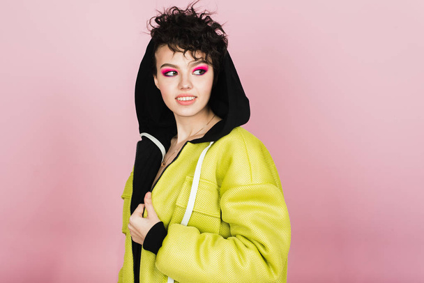 Beautiful teenager girl sincerely smiles and laughs in sports stylish clothes in the studio on a pink background. Bright girl with a short flying hairstyle in a puffed neon lemon jacket and pink smoke - Photo, image