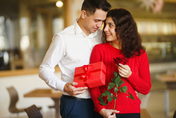 Man gives to his woman a gift box with red ribbon. A loving couple cuddles and celebrating Valentine's Day in the restaurant. Valentine's Day, holiday and surprise concept. Relationship and love. - Foto, afbeelding