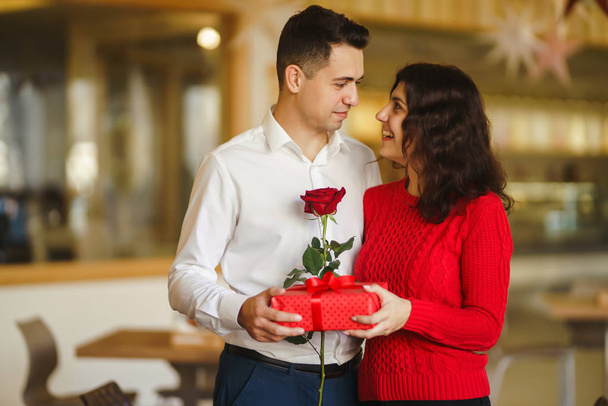 Man gives to his woman a gift box with red ribbon. A loving couple cuddles and celebrating Valentine's Day in the restaurant. Valentine's Day, holiday and surprise concept. Relationship and love. - Photo, image