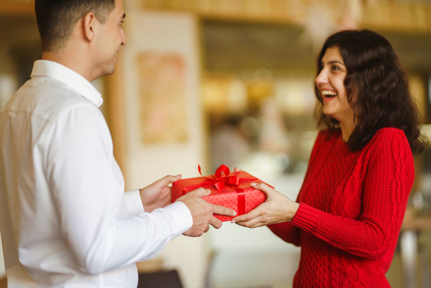 Man gives to his woman a gift box with red ribbon. A loving couple cuddles and celebrating Valentine's Day in the restaurant. Valentine's Day, holiday and surprise concept. Relationship and love. - Photo, image