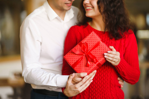 Man gives to his woman a gift box with red ribbon. A loving couple cuddles and celebrating Valentine's Day in the restaurant. Valentine's Day, holiday and surprise concept. Relationship and love. - Φωτογραφία, εικόνα