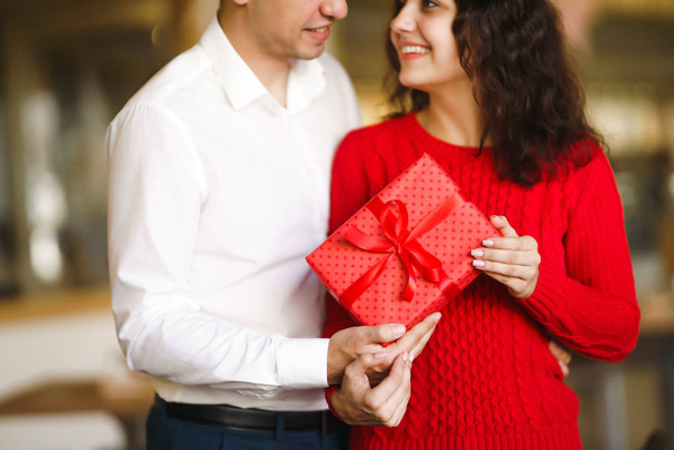 Man gives to his woman a gift box with red ribbon. A loving couple cuddles and celebrating Valentine's Day in the restaurant. Valentine's Day, holiday and surprise concept. Relationship and love. - Фото, изображение