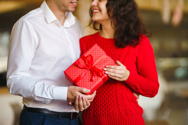 Man gives to his woman a gift box with red ribbon. A loving couple cuddles and celebrating Valentine's Day in the restaurant. Valentine's Day, holiday and surprise concept. Relationship and love. - 写真・画像