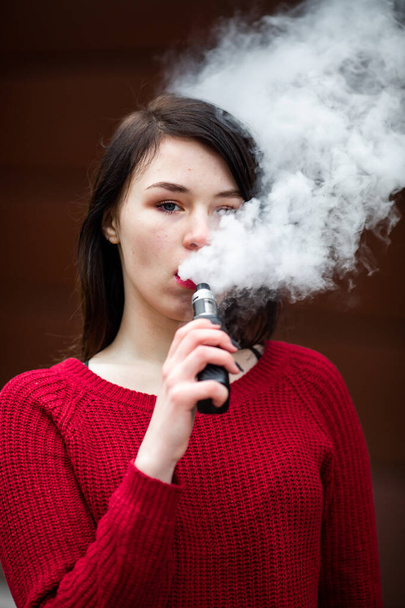 Vape teenager. Young pretty white caucasian girl in a red sweater smoking an electronic cigarette opposite modern brown background on the street in the winter. Deadly bad habit. - Fotoğraf, Görsel