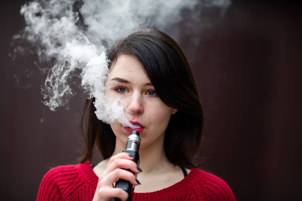 Vape teenager. Young pretty white caucasian girl in a red sweater smoking an electronic cigarette opposite modern brown background on the street in the winter. Deadly bad habit. - 写真・画像