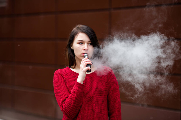 Vape teenager. Young pretty white caucasian girl in a red sweater smoking an electronic cigarette opposite modern brown background on the street in the winter. Deadly bad habit. - Photo, image