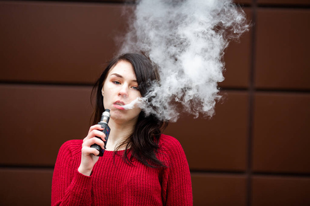 Vape teenager. Young pretty white caucasian girl in a red sweater smoking an electronic cigarette opposite modern brown background on the street in the winter. Deadly bad habit. - Photo, image