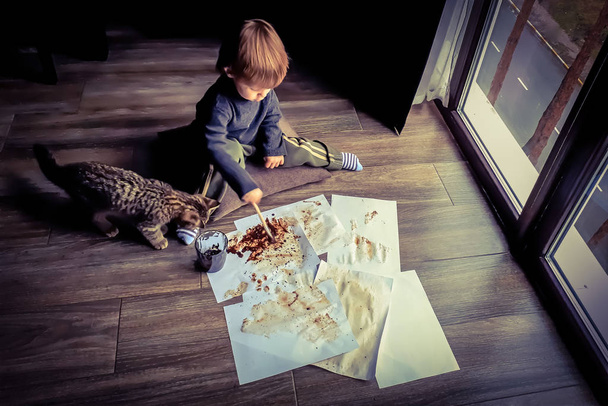 Detailní záběr malého dítěte s pomocnou kočkou, která maluje hnědé kávové sedliny na bílé listy papíru, tmavá podzimní scenérie před velkým oknem doma - Fotografie, Obrázek