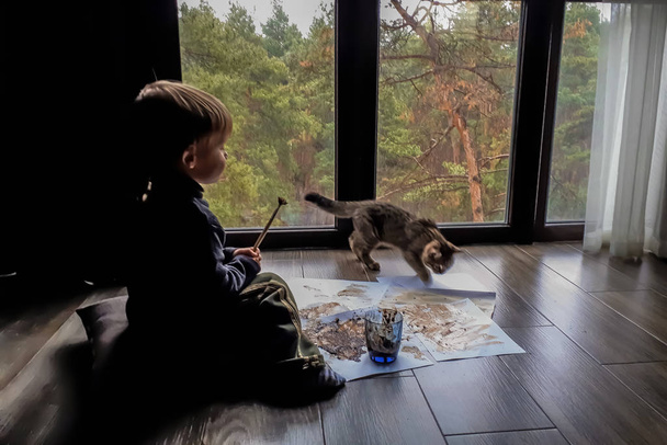 Крупним планом маленька дитина з кішкою-помічником, який малює коричневі кавові майданчики на білих аркушах паперу, темні осінні пейзажі за межами великого вікна вдома
 - Фото, зображення