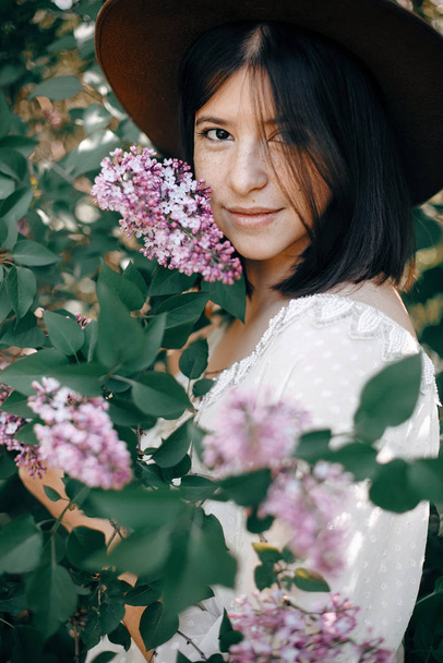 Stylish boho woman in hat posing at lilac flowers in sunny sprin - Foto, imagen