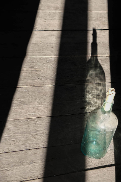 Natura morta rustica con bottiglia di vetro sporco vuota e ombre geometriche su una superficie di legno grezzo. Bottiglia in ragnatela e polvere chiusa con tappo di carta
. - Foto, immagini