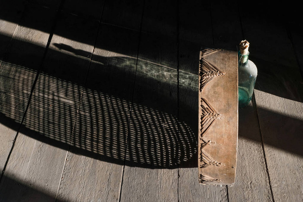  Natura morta rustica con una bottiglia vuota e vecchio setaccio creando belle ombre su una superficie di legno grezzo. Bottiglia in ragnatela e polvere chiusa con tappo di carta
. - Foto, immagini