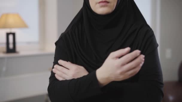 Camera moving up from crossed female hands to face of serious Muslim woman in black hijab looking at camera. Beautiful young lady in posing at home. Eastern people, traditional dress. - Filmagem, Vídeo