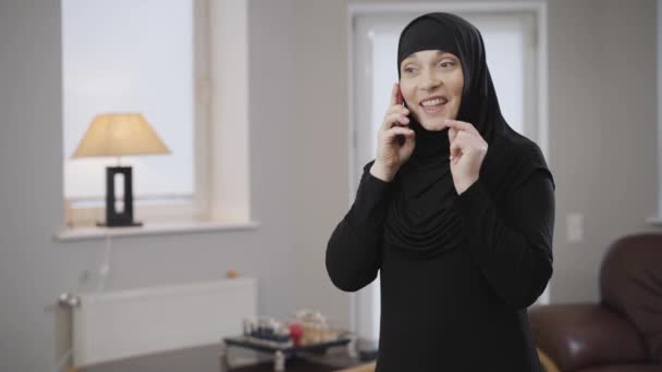 Macchina fotografica si avvicina lentamente alla giovane donna musulmana in hijab parlando su smartphone e sorridendo. Moderna signora orientale utilizzando il telefono cellulare e gesticolando. Tecnologie moderne, cultura tradizionale
. - Filmati, video