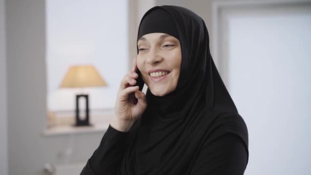 Зблизька молода красива мусульманка в чорному хіджабі з використанням смартфона вдома. Східна леді розмовляє і посміхається. Сучасні технології, традиційна культура. - Кадри, відео