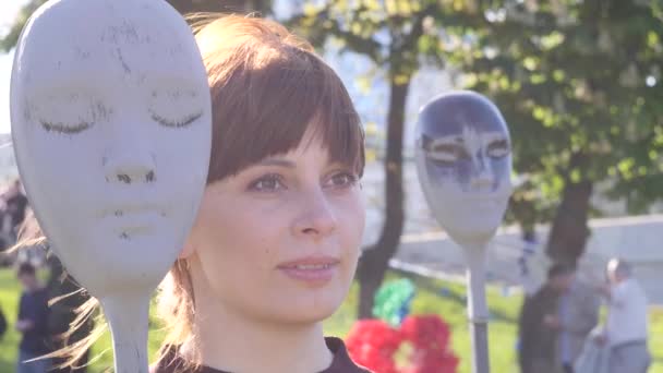 portrait of a young beautiful brunette woman who posing for the camera near the mask of an art installation iagainst the background of the city, close up video in slow motion in 4K - Felvétel, videó