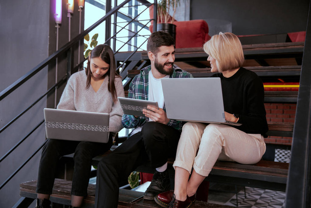 Três jovens trabalhadores de escritório atraentes analisando o plano de trabalho usando o caderno e tablet enquanto estão sentados nas escadas do escritório
 - Foto, Imagem